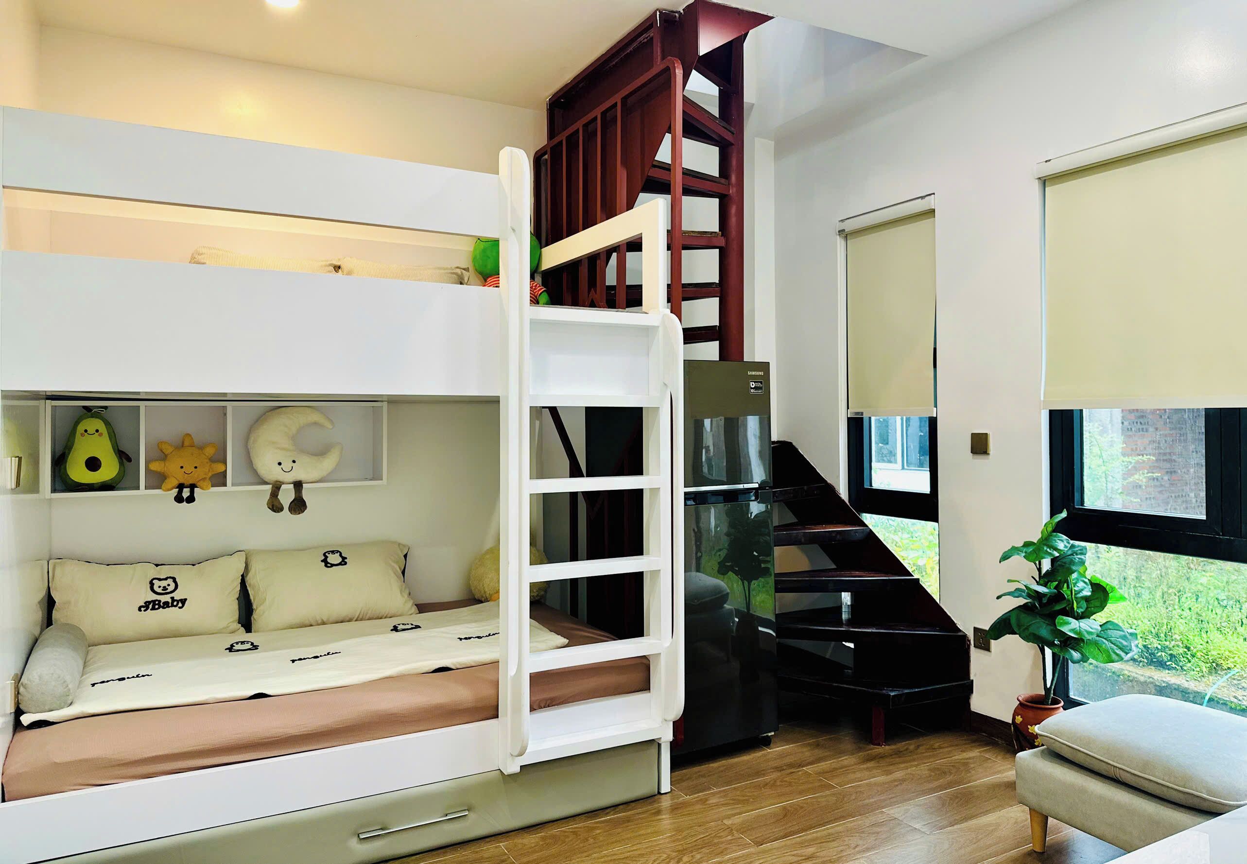 A bunk bed with a wooden ladder, a desk, and a chair. There is a window with a view of greenery.