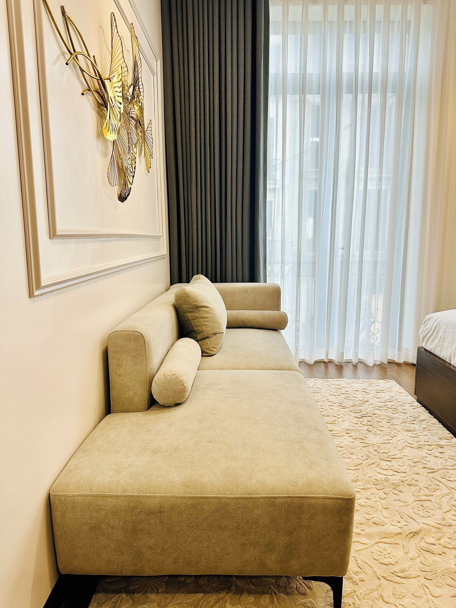 A modern living room with a beige sofa, a decorative wall piece, and a large window with sheer curtains.