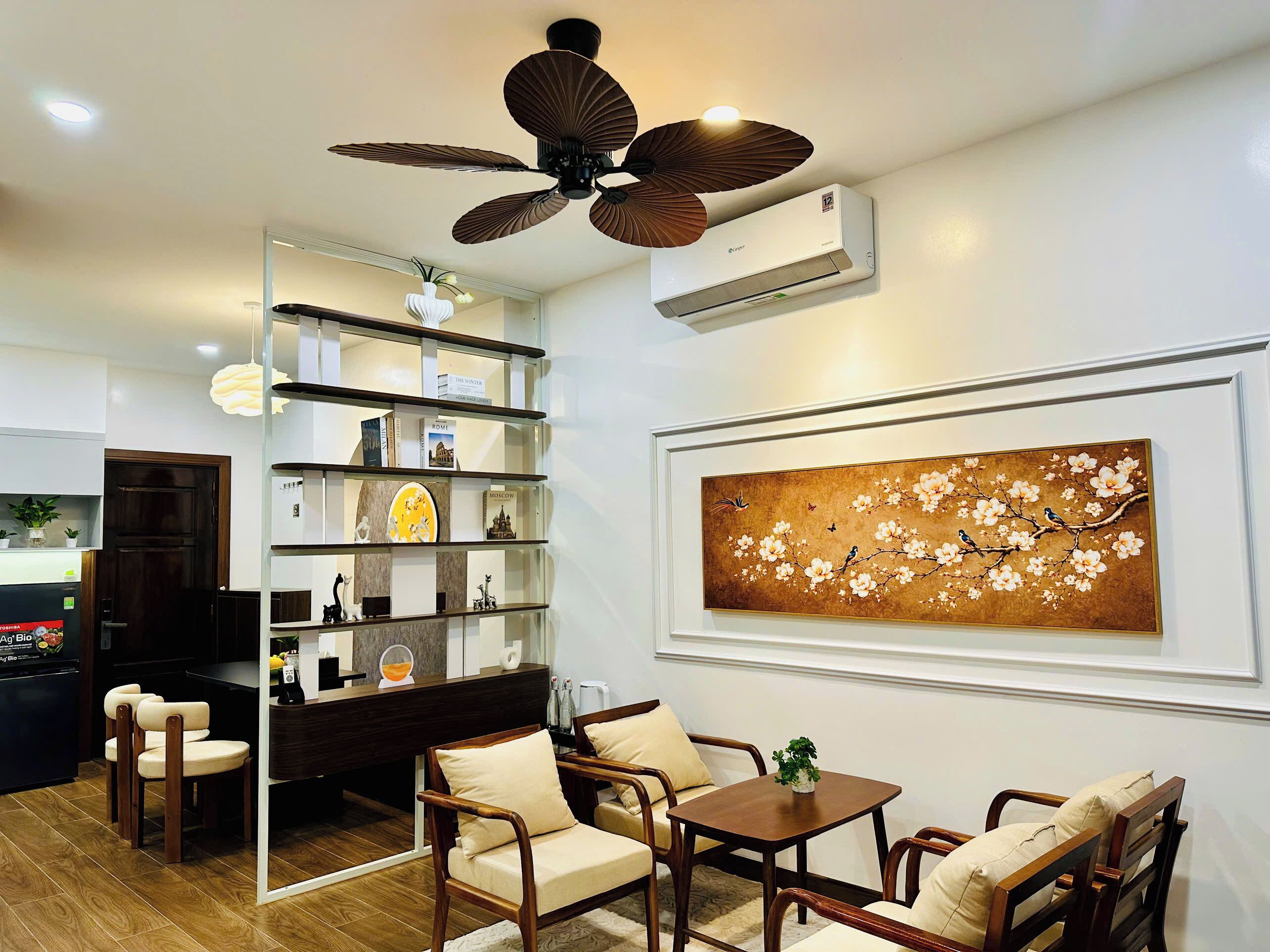 A modern living room with a ceiling fan, a mounted air conditioner, a bookshelf, and a framed artwork on the wall.