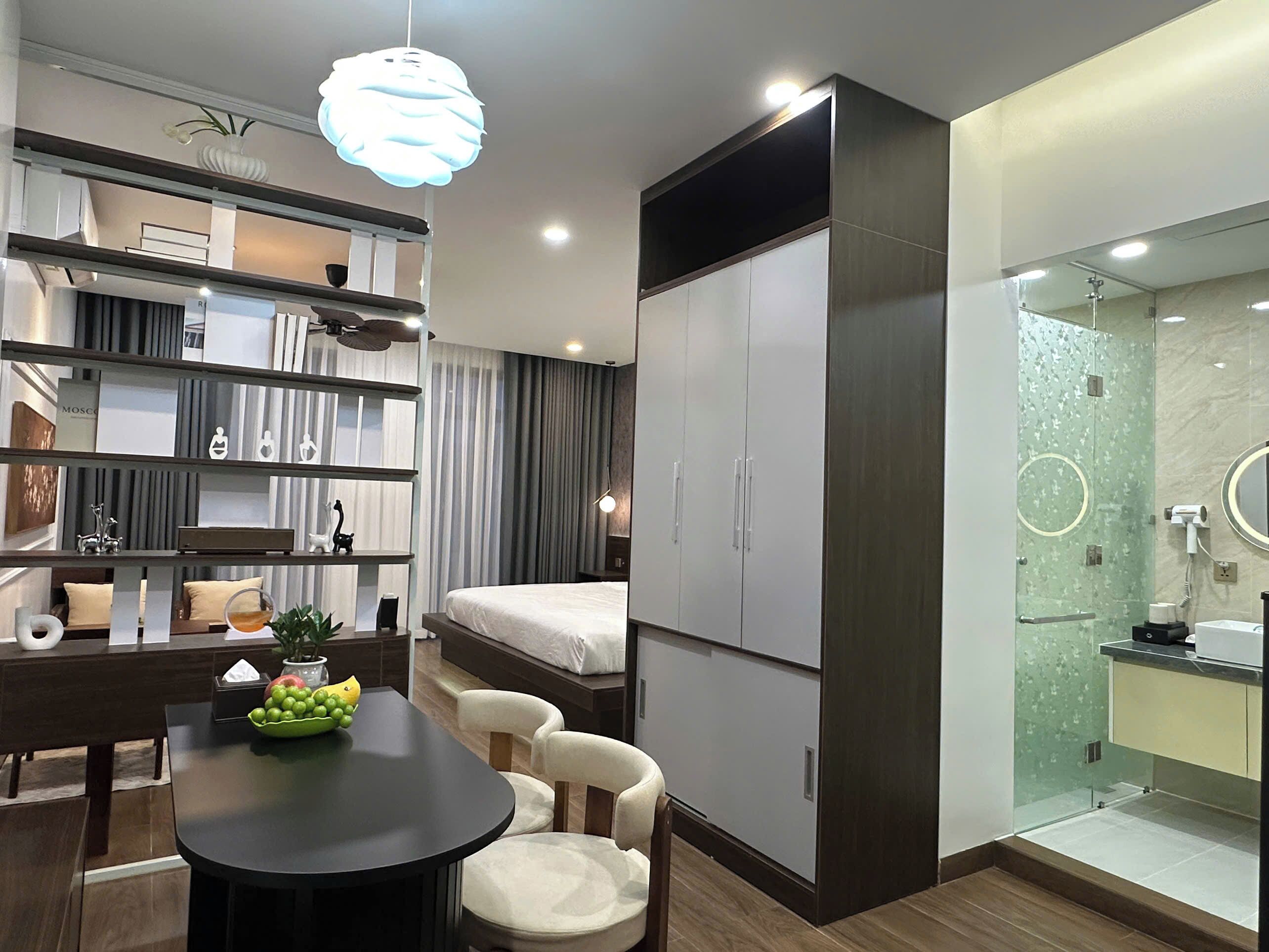 Quarto de hotel moderno com cama, escrivaninha com cadeira, guarda-roupa, banheiro com chuveiro de vidro e área de refeições com mesa e cadeiras.