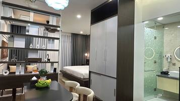 Quarto de hotel moderno com cama, escrivaninha com cadeira, guarda-roupa, banheiro com chuveiro de vidro e área de refeições com mesa e cadeiras.