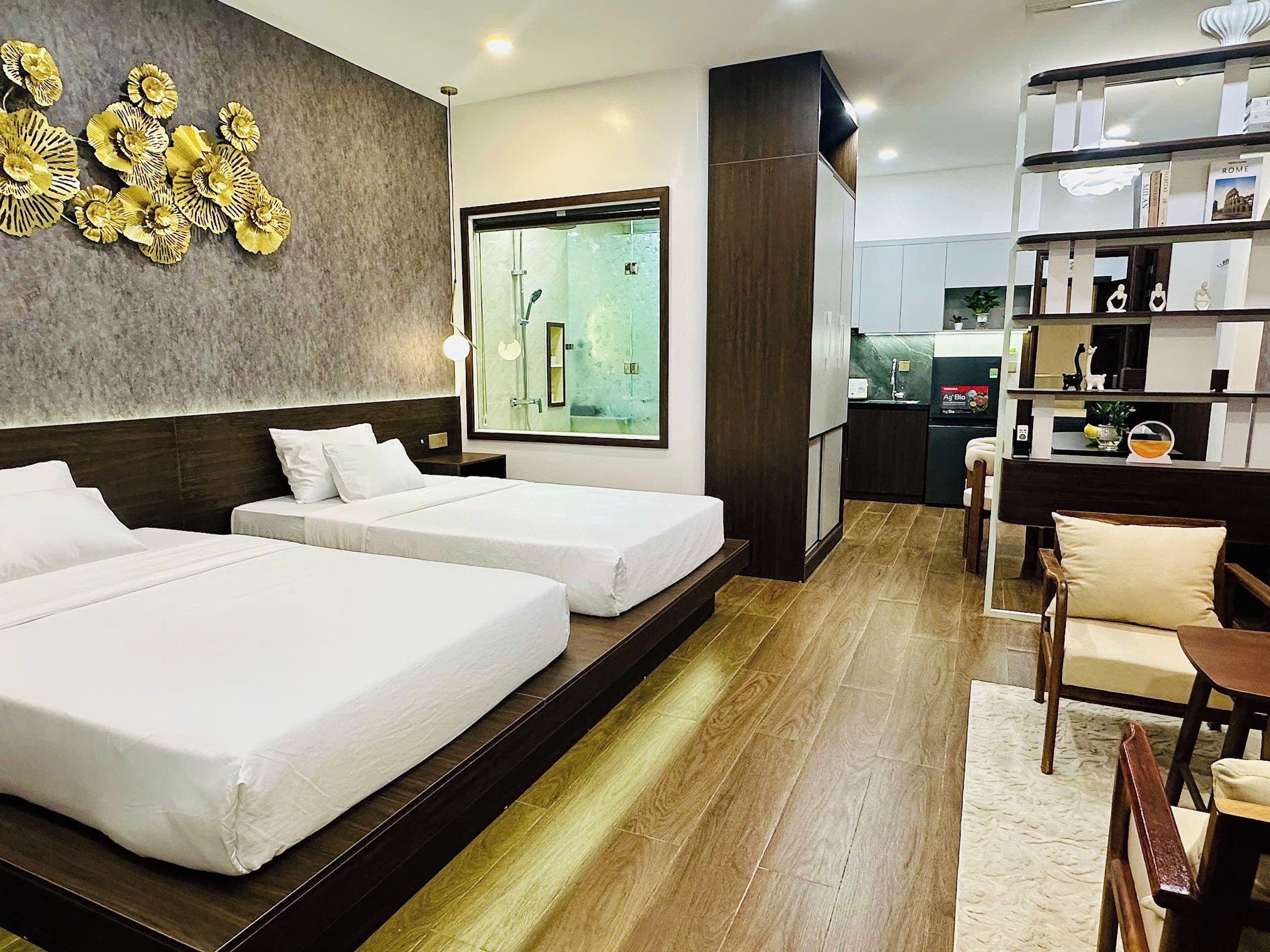 A hotel room with two beds, a large window, a kitchen area, and a bookshelf.