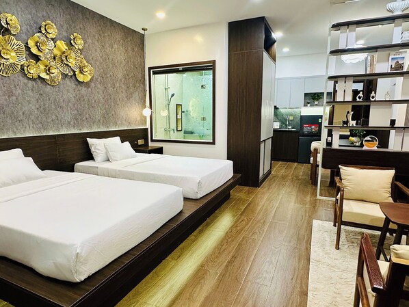 A hotel room with two beds, a large window, a kitchen area, and a bookshelf.