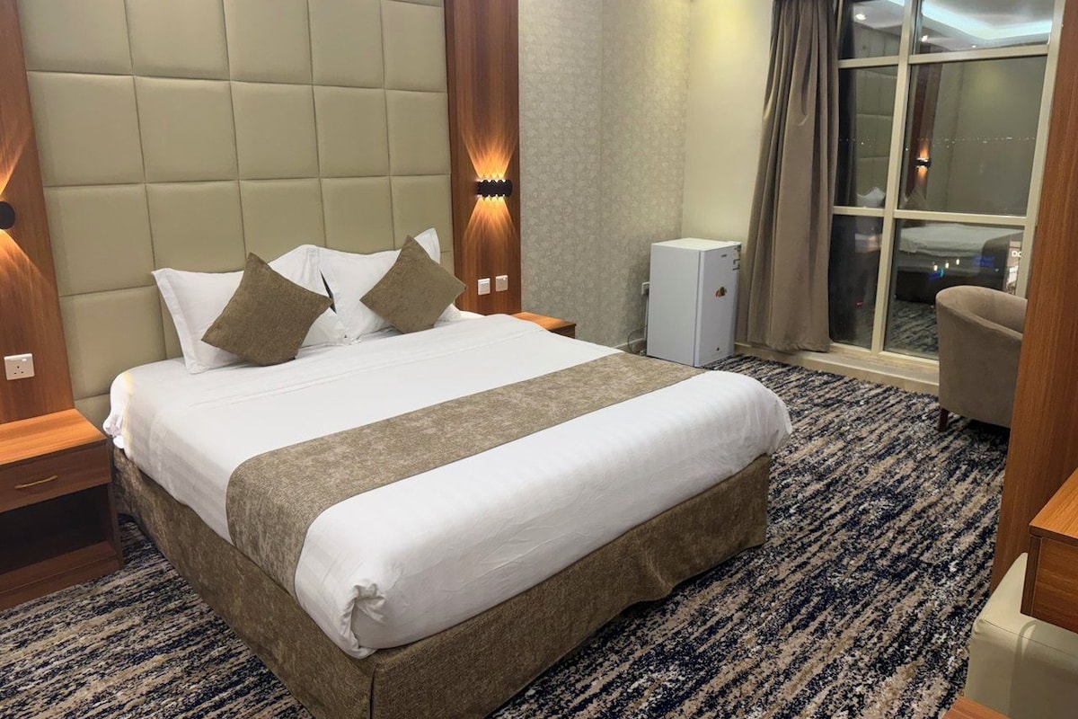 A hotel room with a large bed, a nightstand, a desk, a chair, a wardrobe, and a window with curtains.