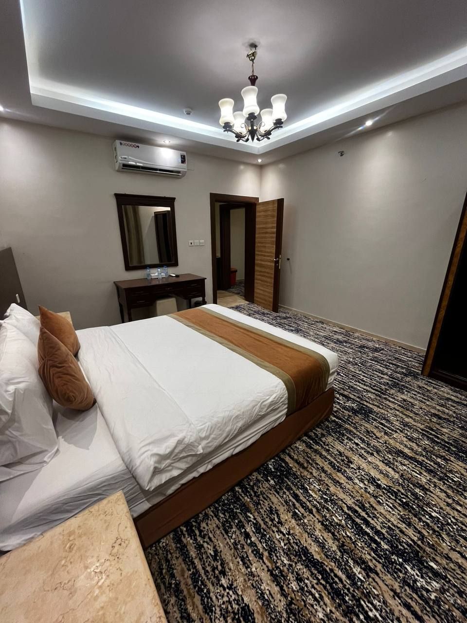 A hotel room with a bed, a desk, a mirror, an air conditioning unit, and a chandelier.