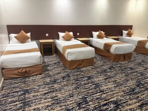 A hotel room with four beds arranged in a row, each with a wooden headboard, white bedding, and brown accents. The room has a patterned carpet and a wooden floor.