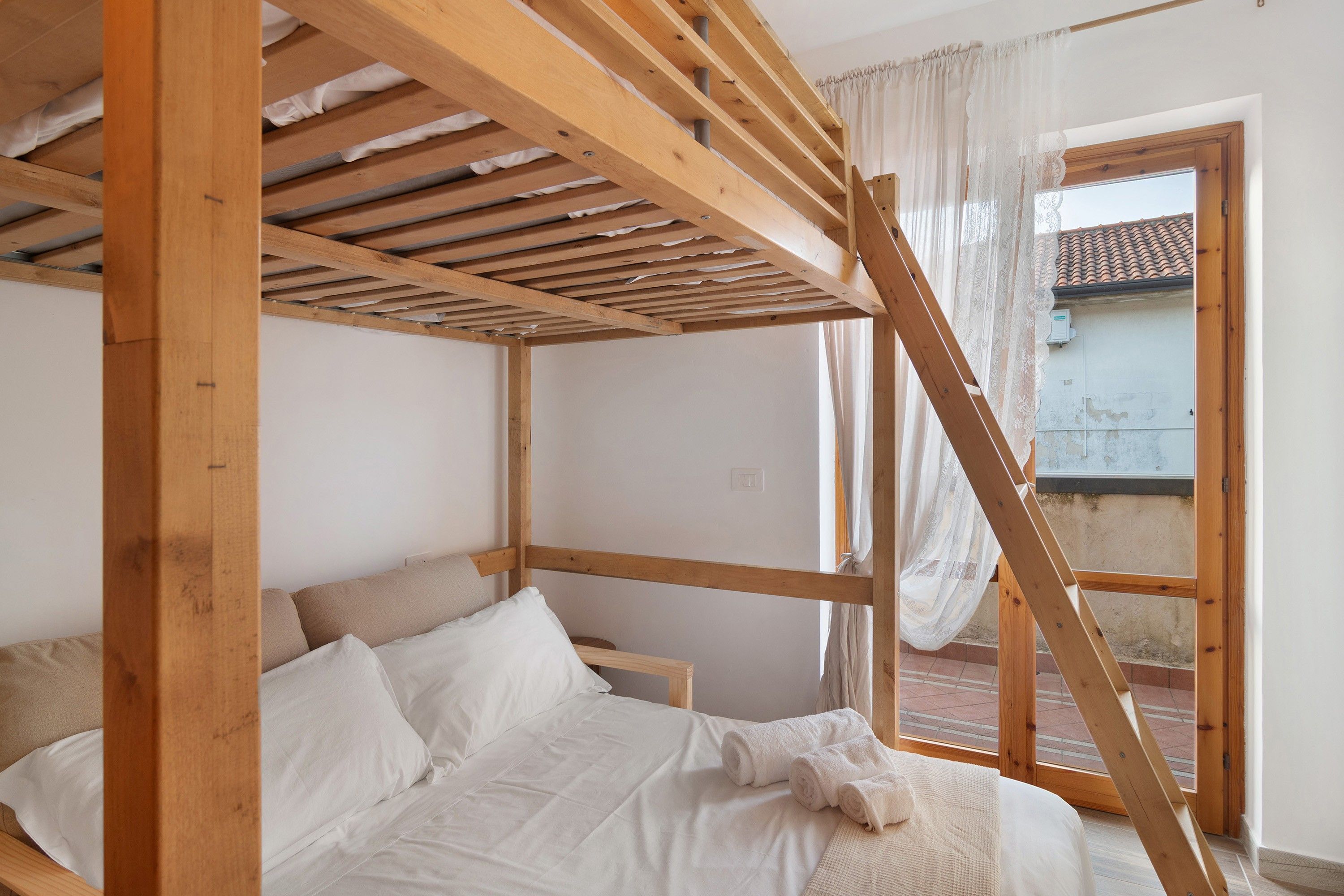 A room with a wooden bunk bed, a window with white curtains, and a view of the outside.