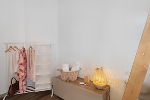 A room with a wooden dresser, a hanging clothes rack, a basket with towels, a lamp, and a mirror.