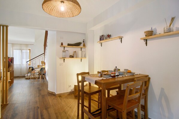 A dining area with a wooden table and chairs, a hanging light fixture, and shelves with various items.
