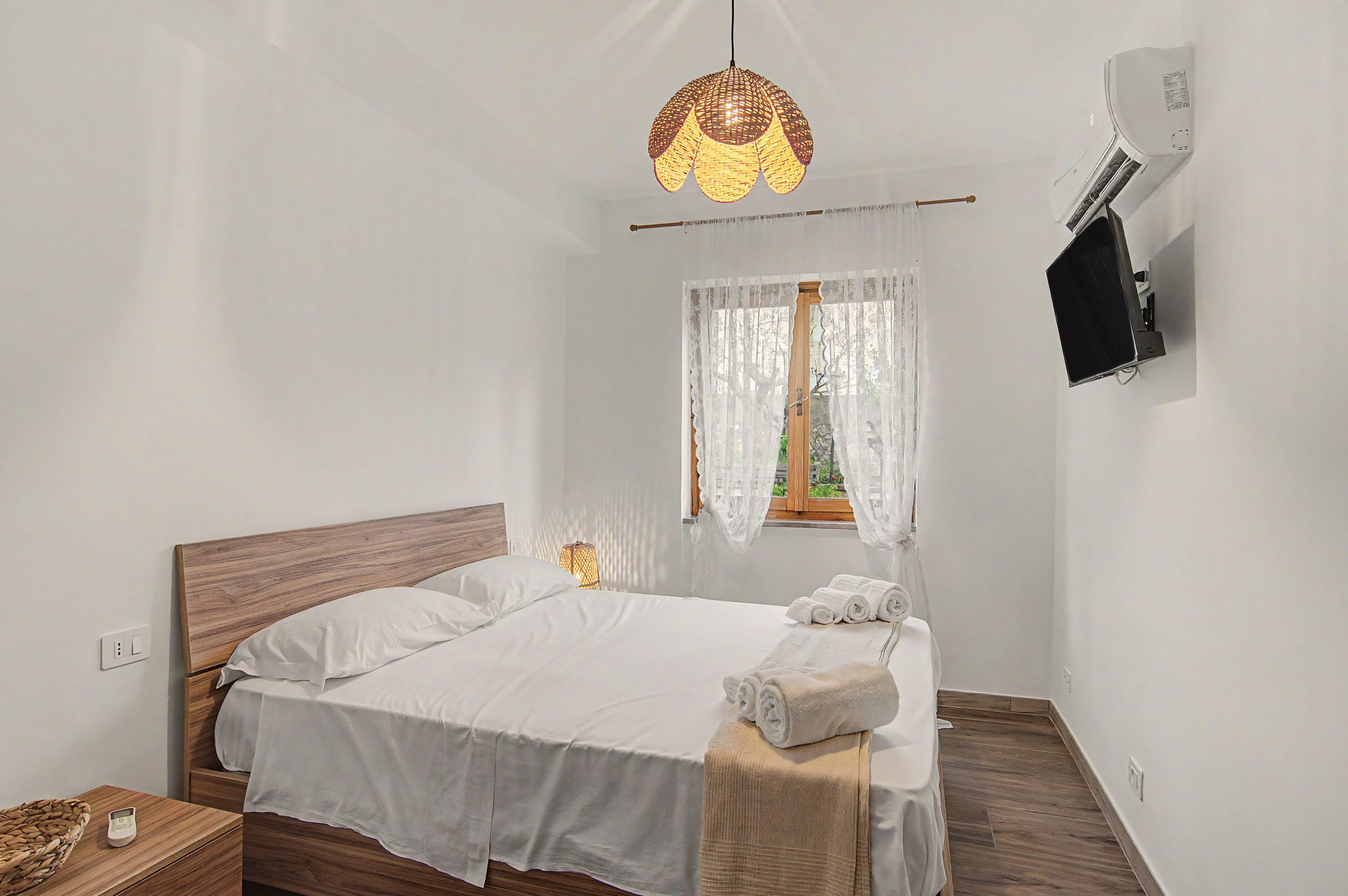 A bedroom with a bed, a bedside table with a lamp, a window with curtains, and an air conditioning unit.