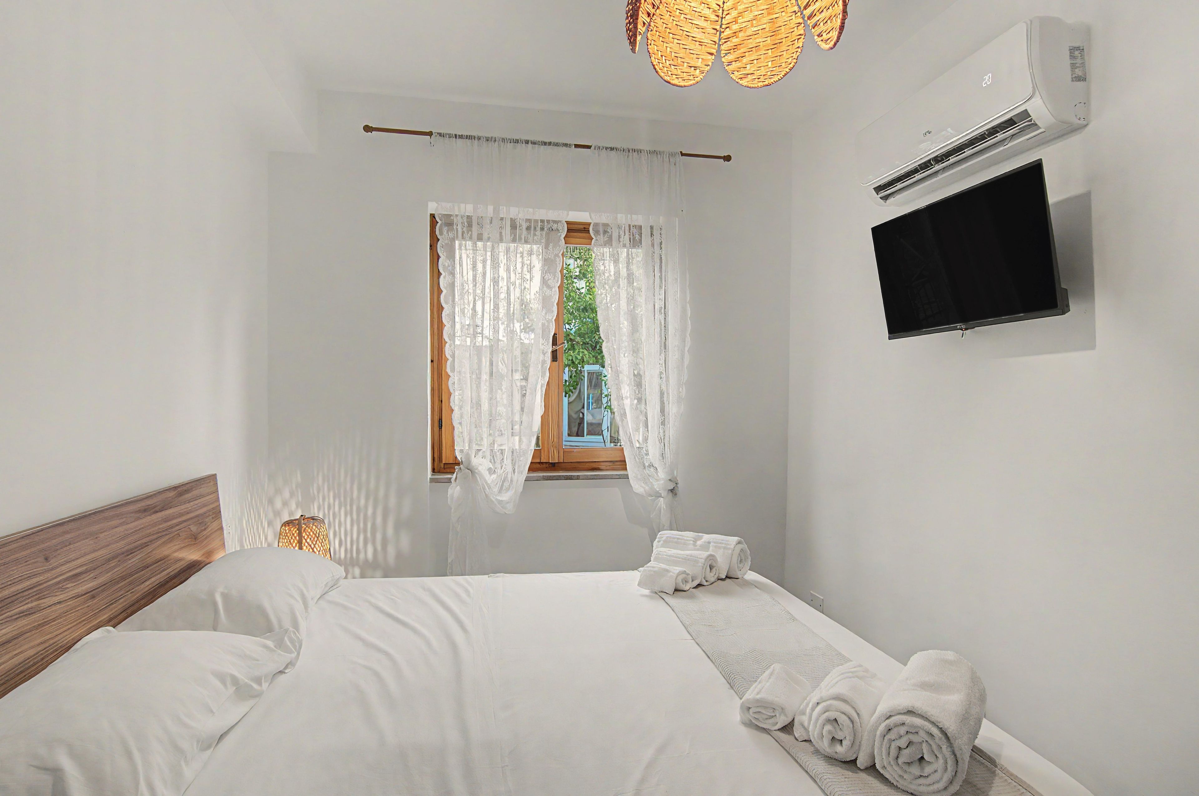 Ein Schlafzimmer mit einem Bett, einem Fenster mit durchsichtigen Vorhängen, einem wandmontierten Fernseher und einem Deckenventilator.