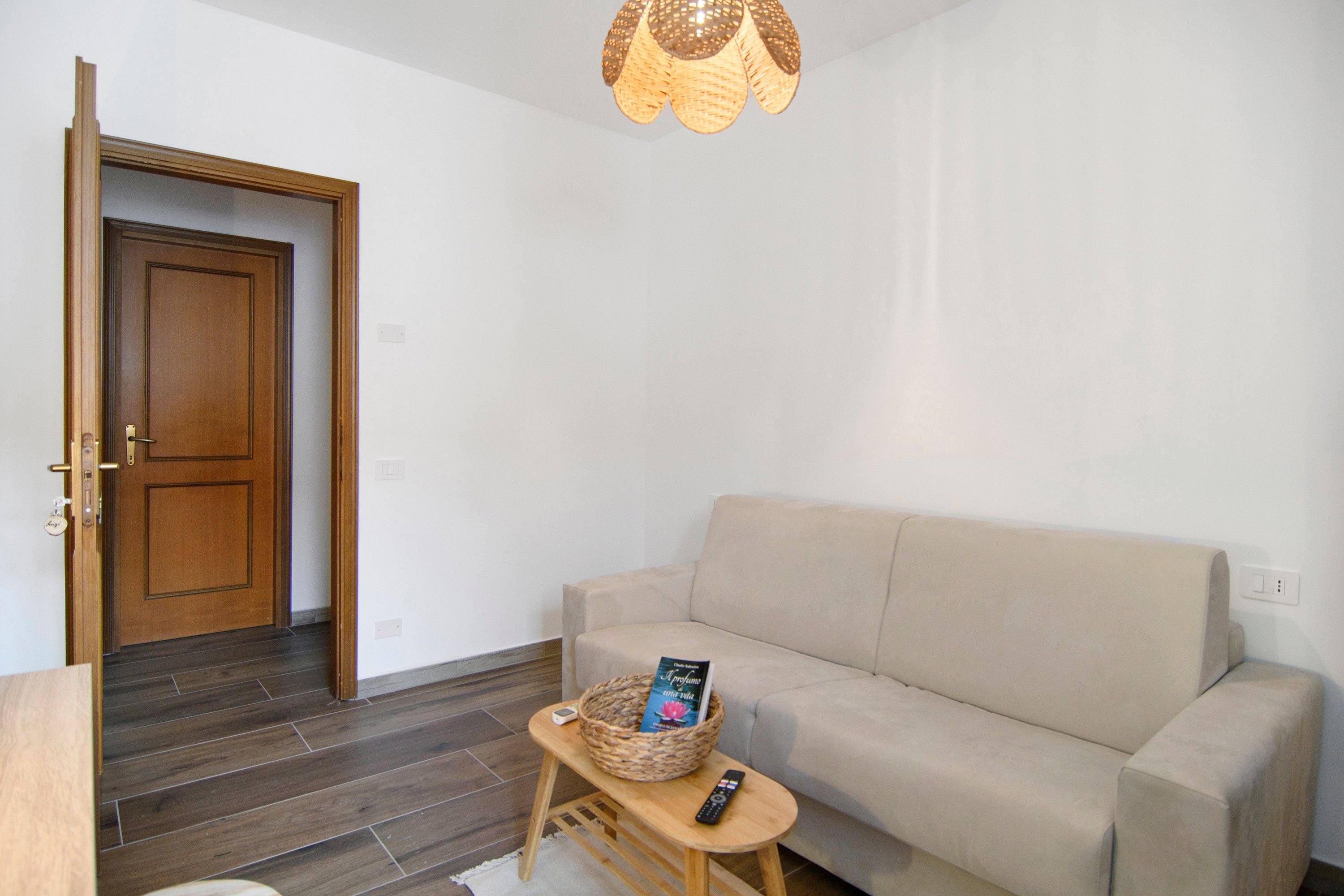 Un salon avec un canapé beige, une table basse en bois, un panier en osier contenant un livre et une télécommande. On aperçoit une porte en bois au fond.