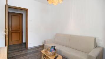 Un salon avec un canapé beige, une table basse en bois, un panier en osier contenant un livre et une télécommande. On aperçoit une porte en bois au fond.