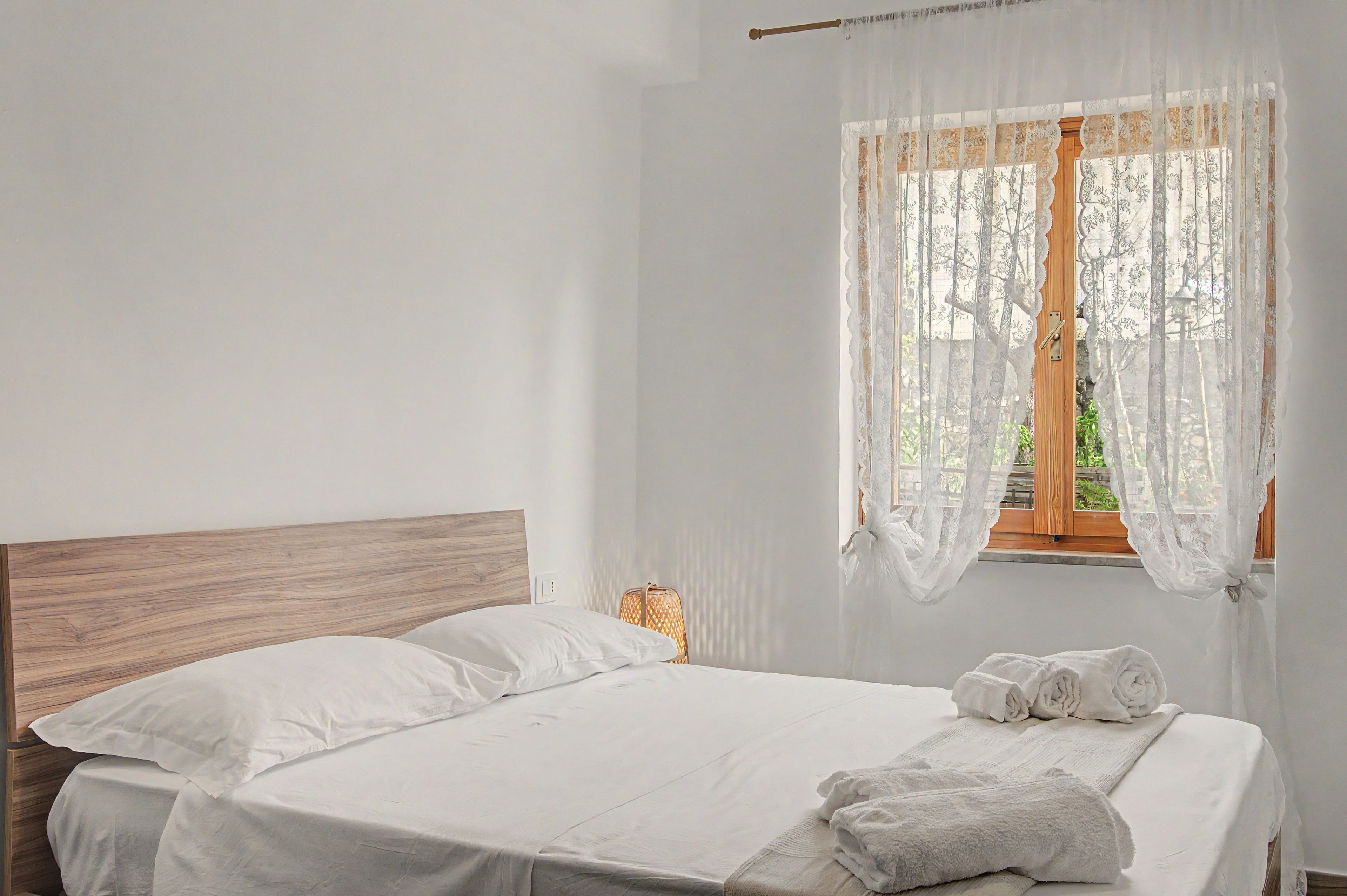 Un lit bien fait, avec du linge de lit blanc et une tête de lit en bois, dans une pièce dotée d’une fenêtre recouverte de rideaux légers.