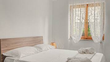 Un lit bien fait, avec du linge de lit blanc et une tête de lit en bois, dans une pièce dotée d’une fenêtre recouverte de rideaux légers.