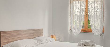 Una cama bien hecha con sábanas blancas y un cabecero de madera en una habitación con cortinas traslúcidas en la ventana.