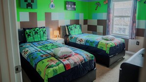 Una camera da letto con due letti, ognuno con copriletto a tema Minecraft, una finestra con tende e una parete decorata con motivi a blocchi di Minecraft.
