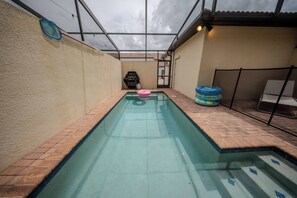 Une piscine rectangulaire avec un bord carrelé, un barbecue et des jouets gonflables.