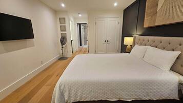 A bedroom with a large bed, a flat-screen TV mounted on the wall, a bedside lamp, and a door leading to another room.