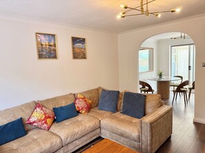 Una sala de estar con un sofá seccional color beige, dos cuadros de paisajes enmarcados, una zona de comedor con una mesa redonda y sillas, y una lámpara colgante.