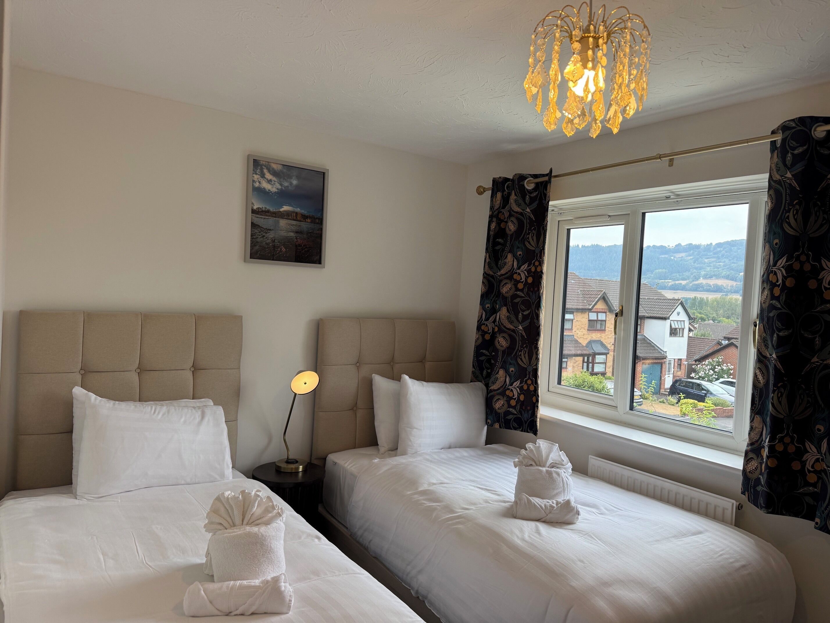 A hotel room with two beds, a bedside table with a lamp, a window with curtains, and a painting on the wall.