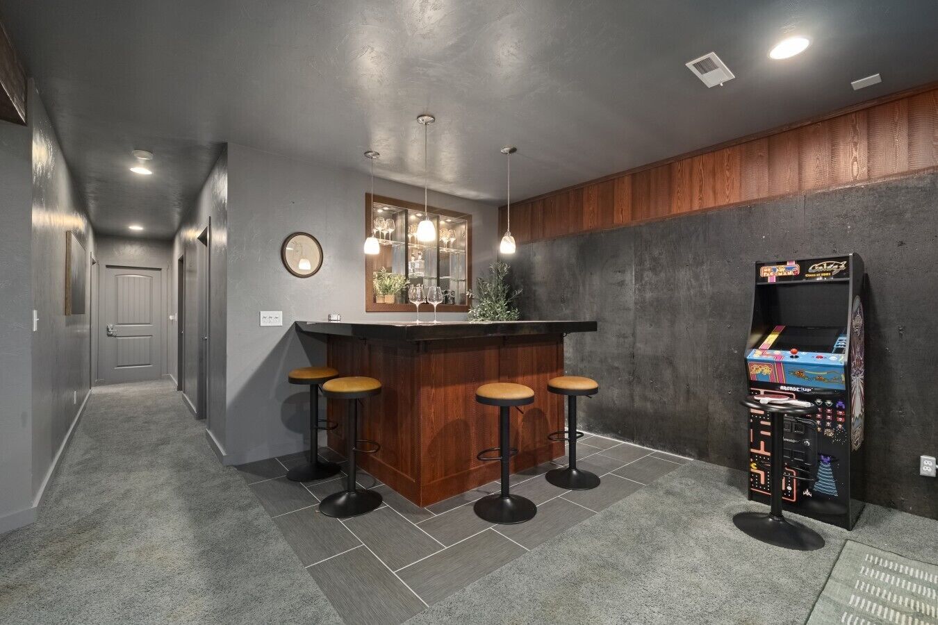 A bar area with a wooden counter, four stools, pendant lighting, a mirror, and a retro arcade machine.