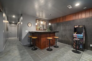 A bar area with a wooden counter, four stools, pendant lighting, a mirror, and a retro arcade machine.