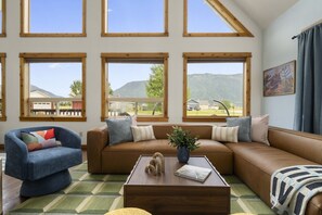 A living room with a brown sofa, a blue armchair, and a wooden coffee table. There are large windows with a view of a mountain and a fence.
