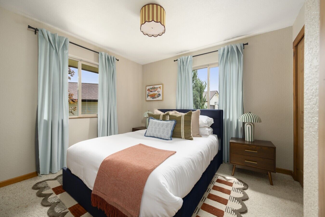 Quarto com cama, mesa de cabeceira, luminária e janela com cortinas.