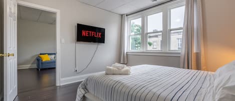 A hotel room with a bed, a TV mounted on the wall, a window with curtains, and a door leading to another room.