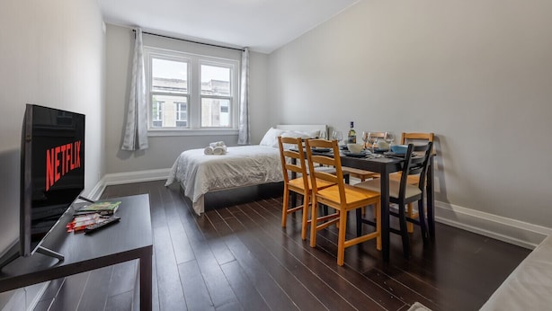 A bedroom with a bed, a TV, a dining table with chairs, and a window with curtains. - Serenity by the Falls: Modern 2 Bedroom Hideaway (Niagara Falls)