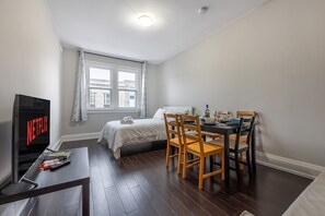 Ein Schlafzimmer mit einem Bett, einem Fernseher, einem Esstisch mit Stühlen und einem Fenster mit Vorhängen.