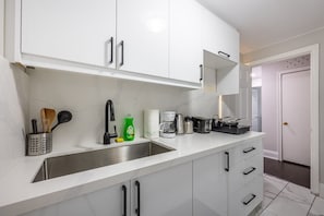A modern kitchen with white cabinets, a stainless steel sink, and various appliances including a coffee maker and toaster. - Serenity by the Falls: Modern 2 Bedroom Hideaway (Niagara Falls)