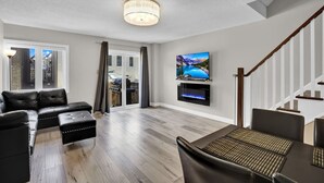 A modern living room with a sectional sofa, a dining table with chairs, a flat-screen TV mounted on the wall, and a staircase with wooden railings. - Stylish Mississauga Lower Unit BBQ Yard Paradise (Mississauga)