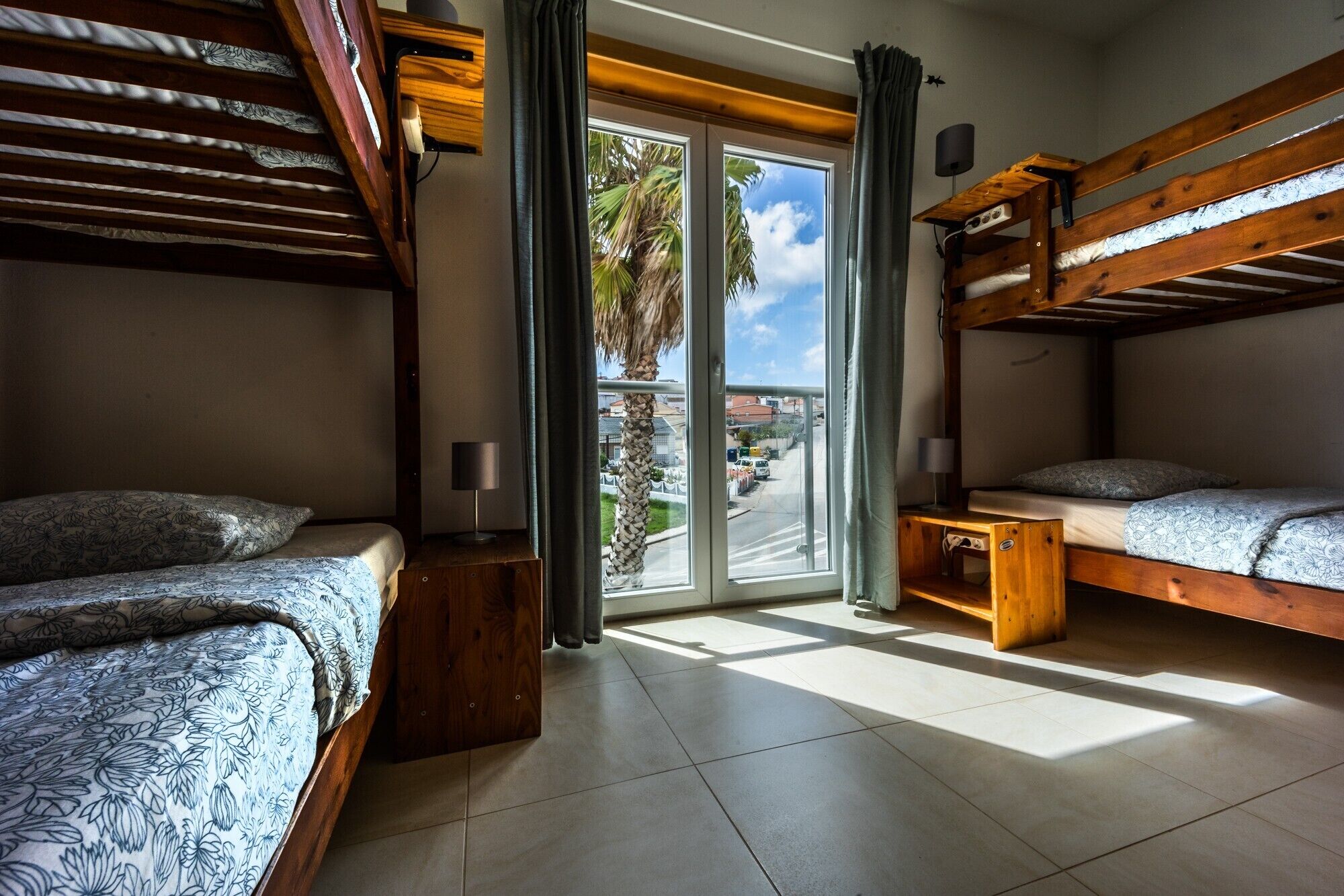A room with two bunk beds, a small table, a lamp, and a view of the street through a glass door.