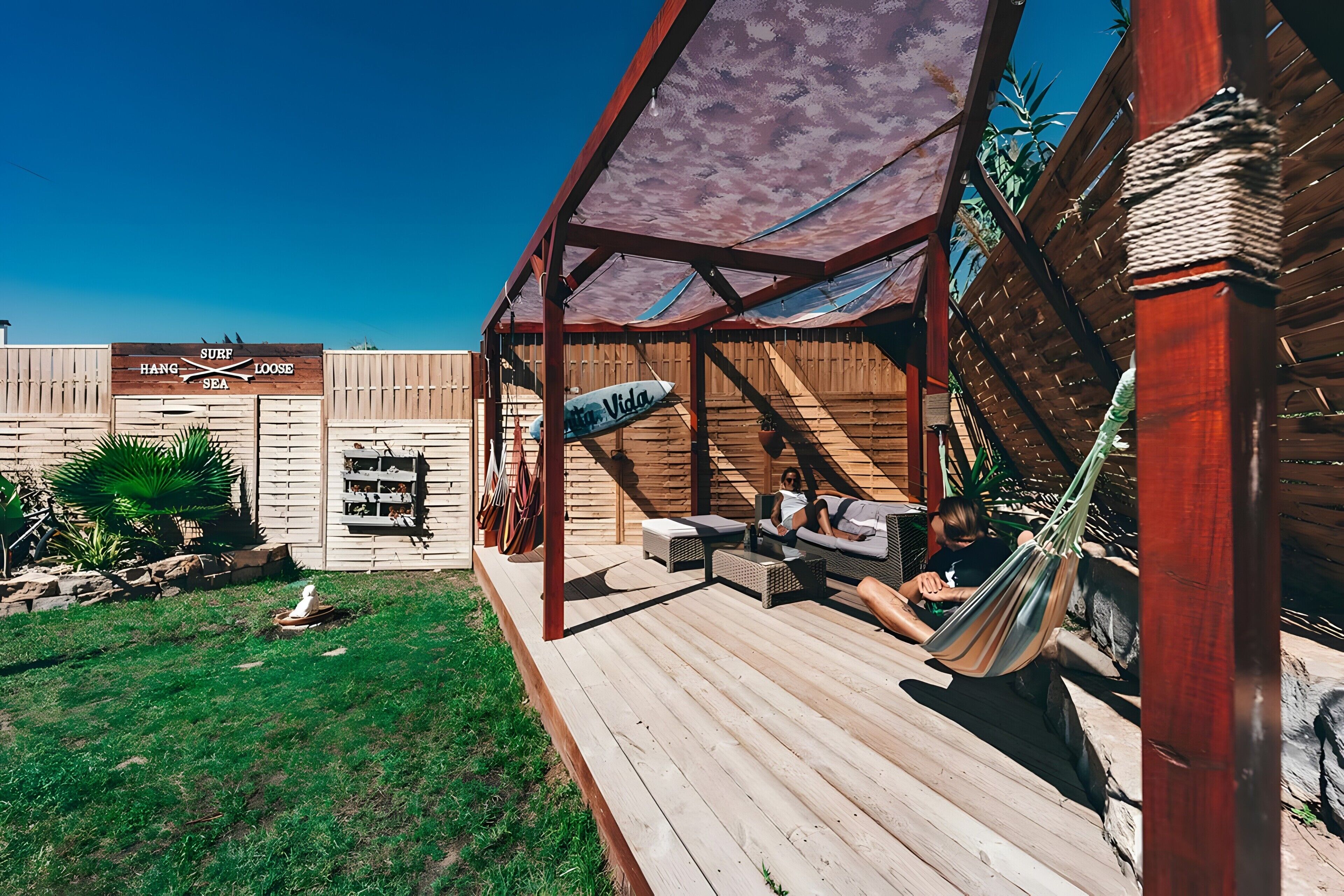 Une terrasse en bois avec des sièges d’extérieur, un hamac et un panneau indiquant « SURF, RELAX, OCEAN ».