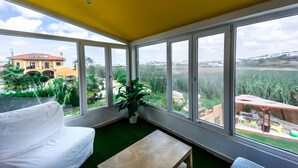 Quarto com um sofá branco, uma mesa pequena e janelas amplas que oferecem vista para um edifício amarelo e áreas verdes.