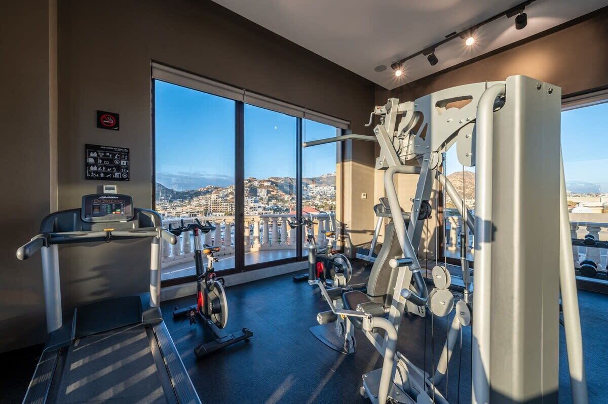 Un gimnasio con equipo cardiovascular y máquinas de pesas, amplias ventanas con vista a la ciudad.