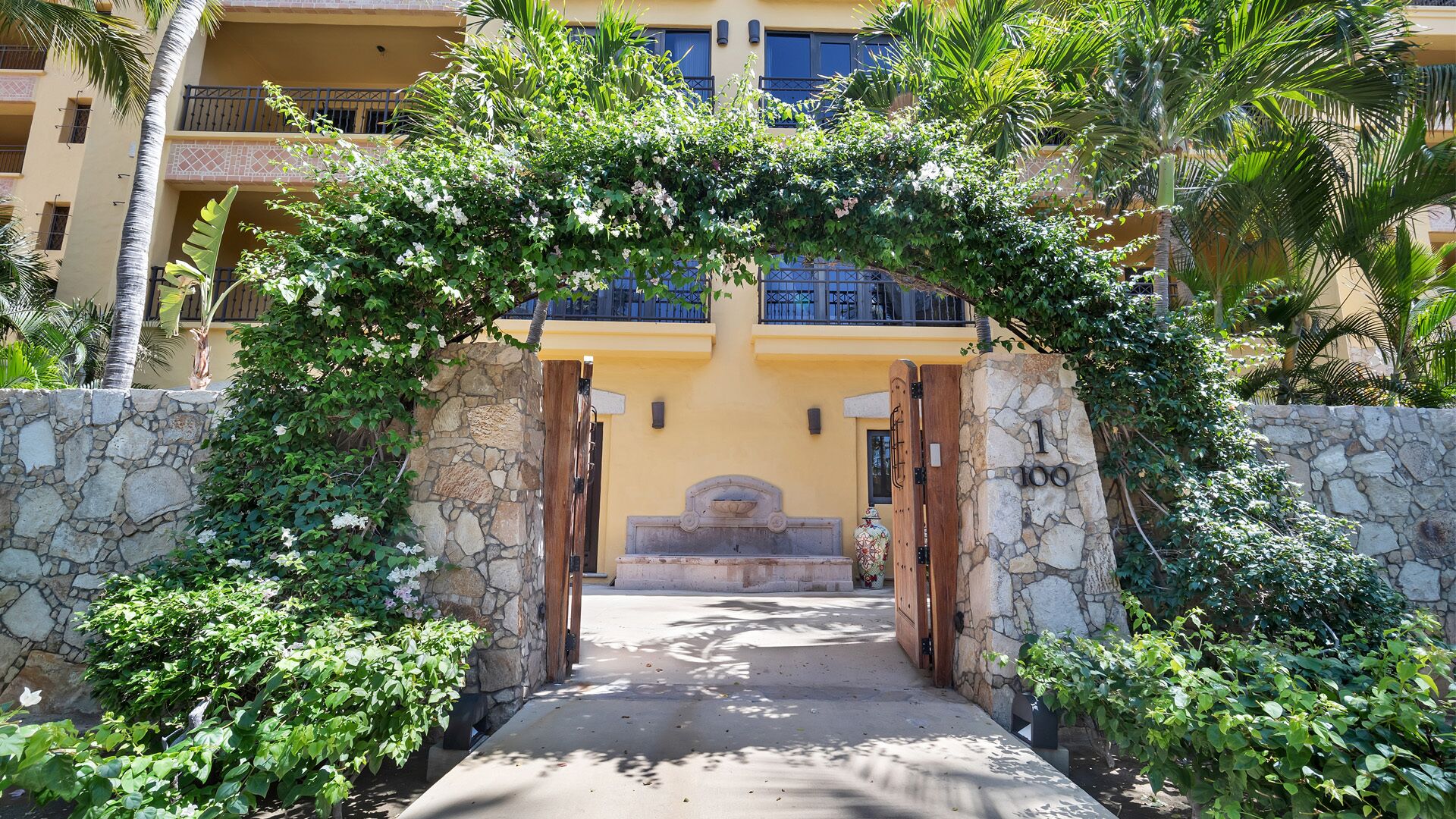 A stone archway leading to a building entrance, surrounded by lush greenery and palm trees.