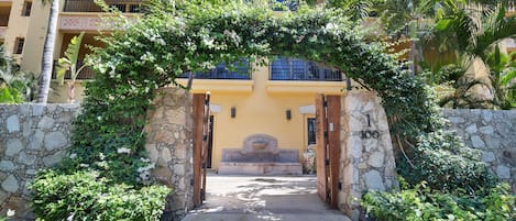 Un arco de piedra que da acceso a la entrada de un edificio, rodeado de exuberante vegetación y palmeras.