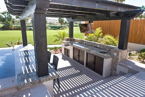 Un espace cuisine extérieur doté d’un plan de travail en pierre, de meubles de rangement intégrés et d’une pergola au-dessus.