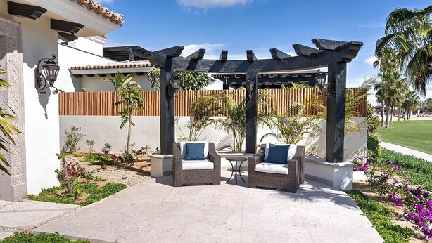 An outdoor patio area with two wicker chairs and a table, surrounded by a wooden pergola and a white wall. - Diamante Golf Villa 39 (Cabo San Lucas)