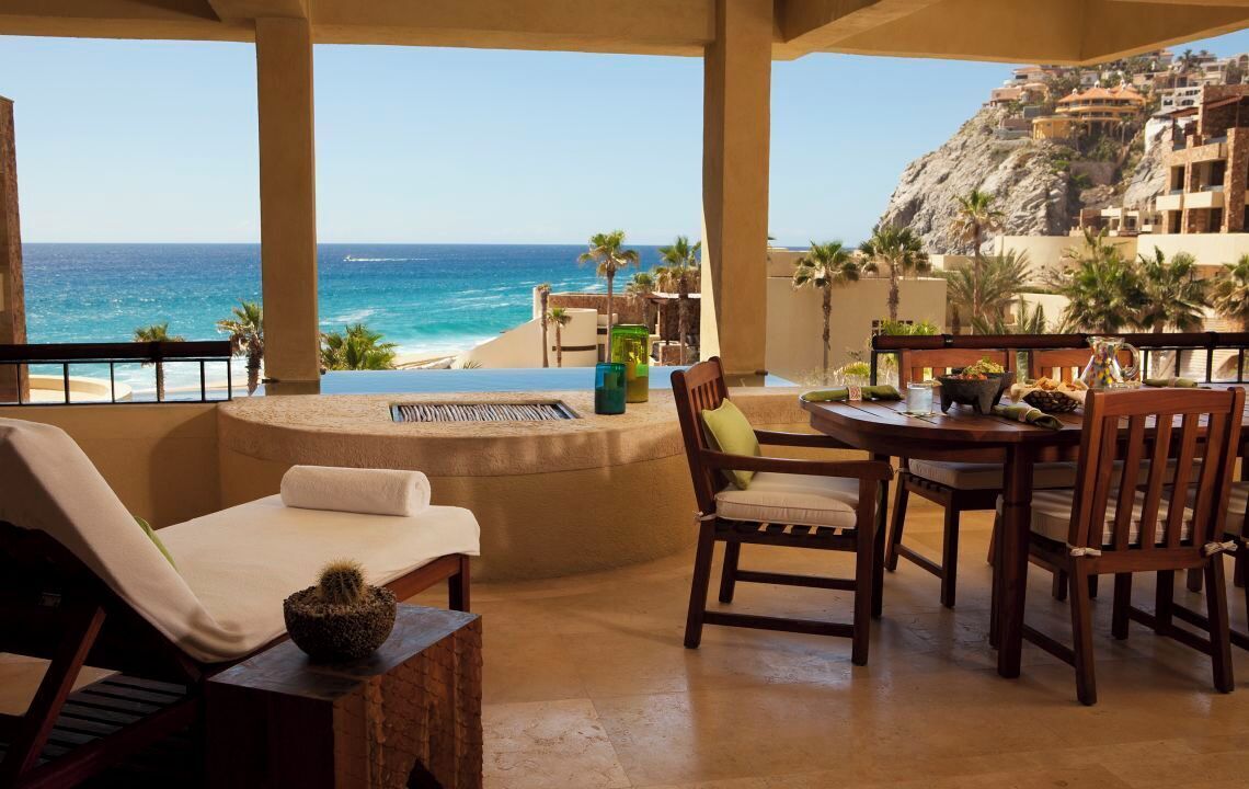 A terrace with a view of the ocean and a mountain, featuring a table and chairs, a lounge chair, and a cactus decoration.