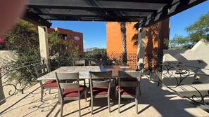 Um pátio externo com mesa e cadeiras, cercado por uma grade de ferro forjado, e edifícios ao fundo.