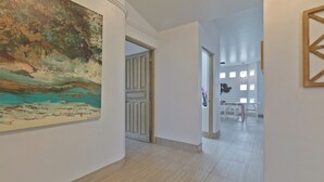 A hallway with a large abstract painting on the left, a door leading to a room with a view, and a dining area in the background. - Marbella lane-Scenic Home with Amazing OceanViews (Cabo San Lucas)
