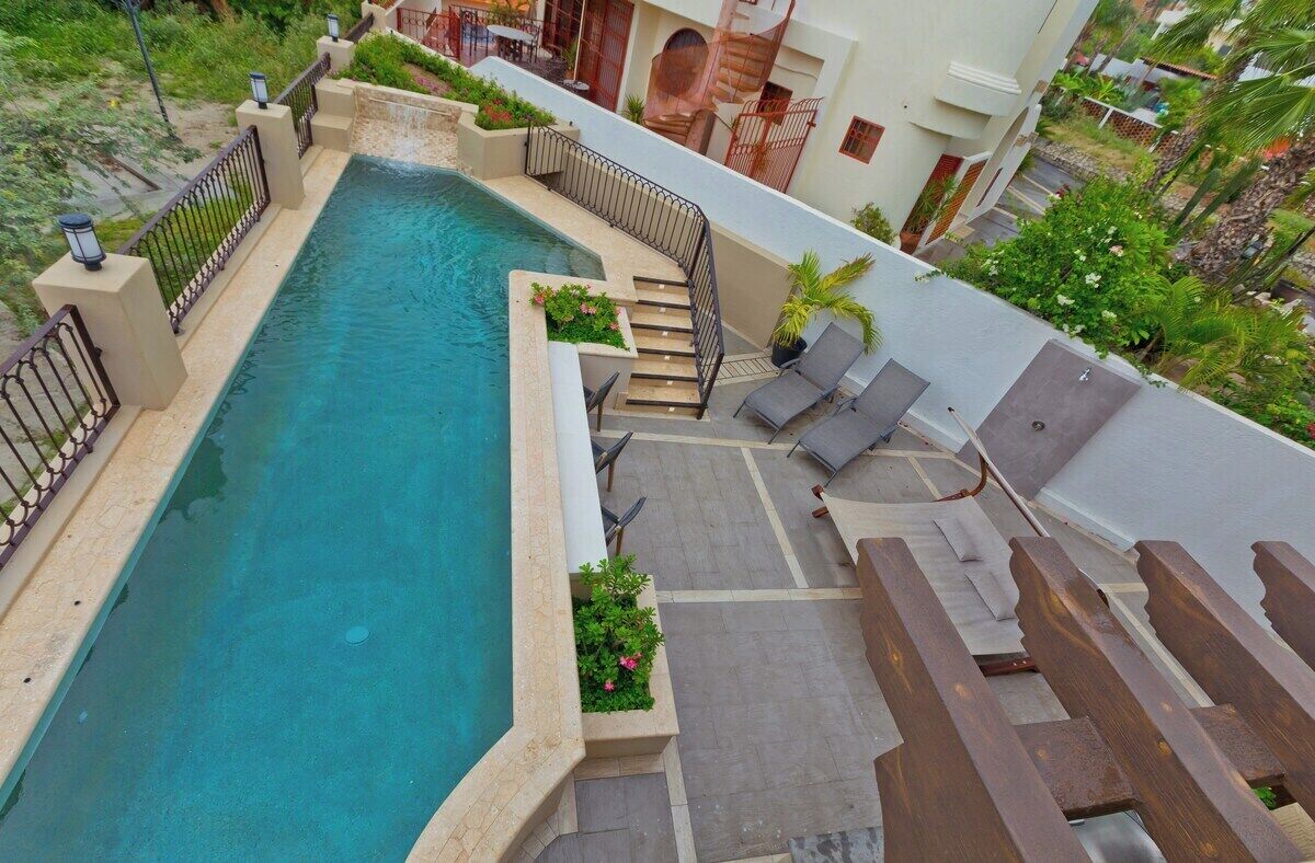 Ein Dachpool mit geschwungener Kante, umgeben von einer niedrigen Mauer und einem Metallgeländer. Treppen führen zum Pool hinab, und einige Liegestühle sind zu sehen.
