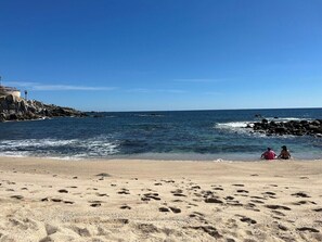 A beach with footprints leading to the water, a rocky outcrop, and two people sitting on the sand. - Marbella Lane PrimeVilla n Modern Home Comfort (Cabo San Lucas)