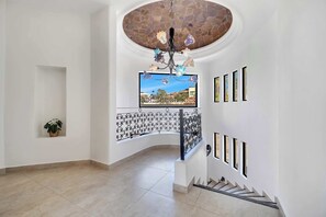 A spacious interior with a stone ceiling, a modern chandelier, a balcony with a wrought iron railing, and a large window offering a view of the outside. - Marbella Lane PrimeVilla n Modern Home Comfort (Cabo San Lucas)