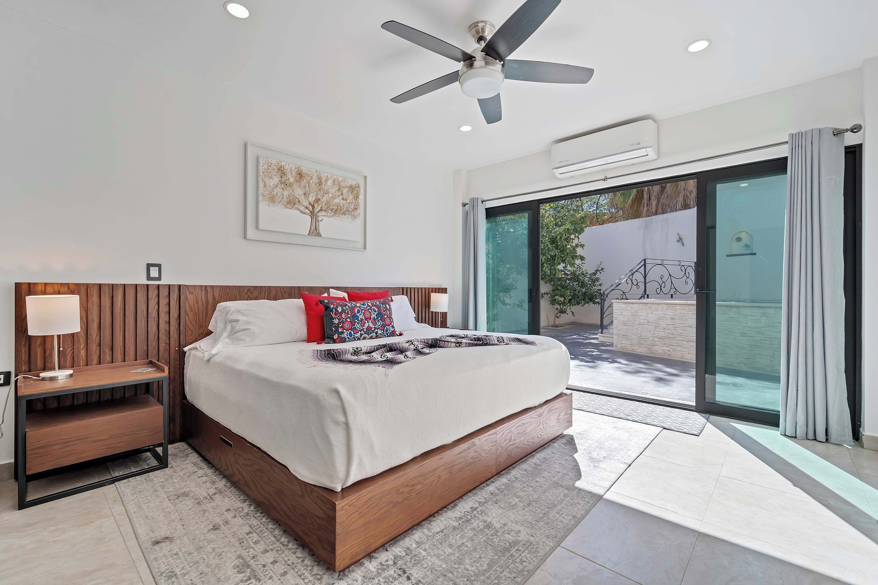 A modern bedroom with a large bed, a ceiling fan, an air conditioning unit, a nightstand, and a view through a glass door to an outdoor area.