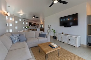 A modern living room with a sofa, coffee table, and TV. There is a kitchen in the background with a ceiling fan. - Marbella Lane Premium Ocean View Villa & Pool (Cabo San Lucas)